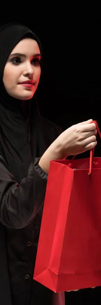 Πορτρέτο του όμορφη έξυπνη νεαρή γυναίκα μουσουλμανικής φορώντας μαύρο χιτζάμπ προσφέροντας τσάντα για ψώνια κατάστημα βοηθός σε μαύρο φόντο — Φωτογραφία Αρχείου