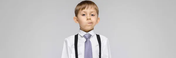 Un niño encantador con una camisa blanca, tirantes, corbata y pantalones vaqueros ligeros se levanta sobre un fondo gris. el chico se torció la cara — Foto de Stock