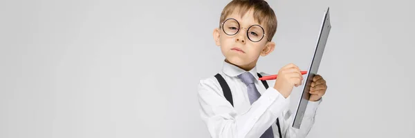 Ein charmanter Junge in weißem Hemd, Hosenträgern, Krawatte und heller Jeans steht vor grauem Hintergrund. der Junge hält einen Stift und Blätter für Notizen — Stockfoto