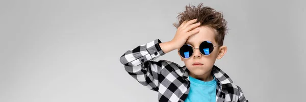 Um menino bonito em uma camisa xadrez, camisa azul e jeans fica em um fundo cinza. O rapaz usa óculos redondos. Rapaz ruivo segura a mão atrás da cabeça — Fotografia de Stock