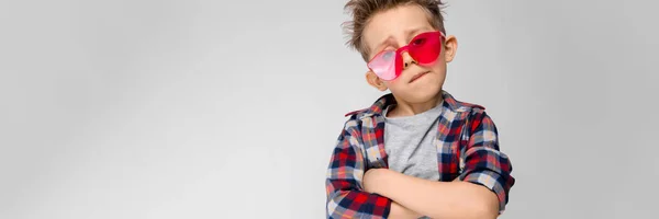 Een knappe jongen in een plaid shirt, grijs shirt en spijkerbroek staat op een grijze achtergrond. Een jongen in rode zonnebril. De jongen zijn armen over zijn borst gevouwen. — Stockfoto