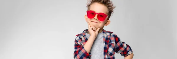 Um menino bonito em uma camisa xadrez, camisa cinza e jeans fica em um fundo cinza. Um rapaz de óculos de sol vermelhos. O menino apoiou o queixo com dois dedos — Fotografia de Stock