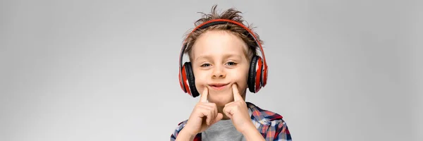 Um menino bonito em uma camisa xadrez, camisa cinza e jeans fica em um fundo cinza. Um rapaz com auscultadores vermelhos. O menino estende os dedos com um sorriso . — Fotografia de Stock