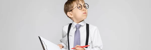 Un niño encantador con una camisa blanca, tirantes, corbata y pantalones vaqueros ligeros se levanta sobre un fondo gris. El chico sostiene un bolígrafo y sábanas para notas —  Fotos de Stock