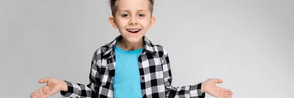 Yakışıklı bir çocuk bir ekose gömlek, mavi tişört ve kot gri bir arka plan üzerinde duruyor. Çocuk elleri her iki yönde yaymak — Stok fotoğraf