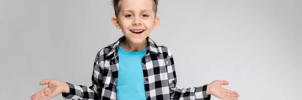 Ein gutaussehender Junge in kariertem Hemd, blauem Hemd und Jeans steht vor grauem Hintergrund. der Junge breitete seine Hände in beide Richtungen aus — Stockfoto