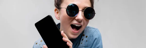 Young brunette girl in round glasses. Hairs are gathered in a bun. Girl with a black phone. — Stock Photo, Image