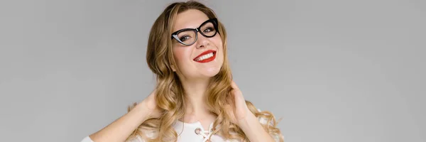 Atractiva joven mujer de negocios rubia en ropa blanca y negra sonriendo sosteniendo su cabello de pie en la oficina sobre fondo gris —  Fotos de Stock