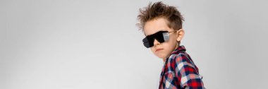 Yakışıklı bir çocuk bir ekose gömlek, gri gömlek ve pantolon gri bir arka plan üzerinde duruyor. Siyah güneş gözlüklü çocuk. Yarı uyanık çocuktur. Çocuk onun kalçalarına ellerini koymak.