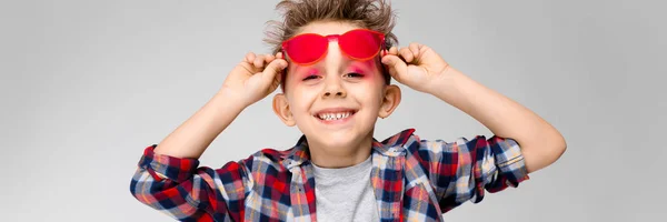 Un chico guapo con una camisa a cuadros, camisa gris y jeans está parado sobre un fondo gris. Un chico con gafas de sol rojas. El chico sostiene sus gafas de mano — Foto de Stock