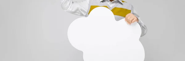 Een charmante jongen in lichte jeans, een wit overhemd en gestreepte tank top staat op een grijze achtergrond. De jongen houdt een witte poster in de vorm van een wolk — Stockfoto