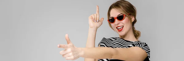 Attraktive junge blonde Mädchen in gestreifter Bluse lächelnd in Sonnenbrille narren stehend auf grauem Hintergrund — Stockfoto
