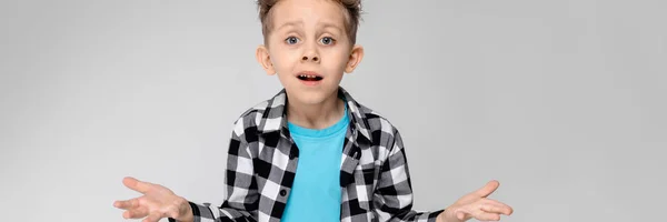 Un chico guapo con camisa a cuadros, camisa azul y pantalones vaqueros está parado sobre un fondo gris. El chico extendió sus manos en ambas direcciones. —  Fotos de Stock