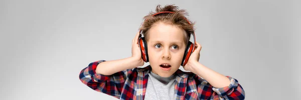 Een knappe jongen in een plaid shirt, grijs shirt en spijkerbroek staat op een grijze achtergrond. Een jongen in rode hoofdtelefoon. De jongen houdt zijn hoofdtelefoon. — Stockfoto