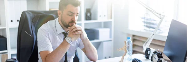 A man is sitting at a table in the office and is looking at a wooden little man. — Stock Photo, Image