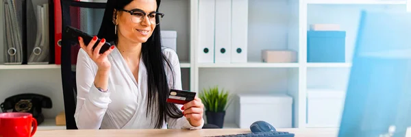 Una joven está sentada a la mesa, sosteniendo una tarjeta bancaria y un teléfono . — Foto de Stock