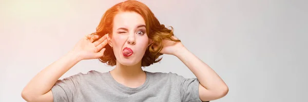 Charming young girl in a gray T-shirt on a gray background. Girl showing tongue — Stock Photo, Image