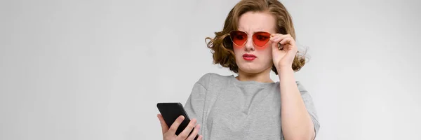 Menina encantadora em uma camiseta cinza em um fundo cinza. Menina em óculos vermelhos com telefone — Fotografia de Stock