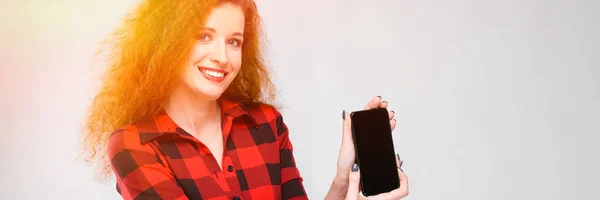 Menina ruiva jovem em uma camisa quadriculada vermelha. Menina mostra telefone — Fotografia de Stock