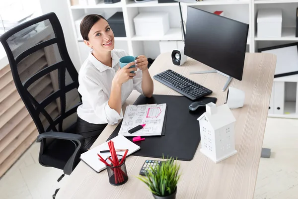 Een jong meisje zittend op kantoor op de computer en holding een kop. — Stockfoto