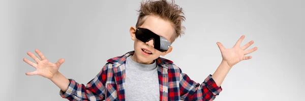 A handsome boy in a plaid shirt, gray shirt and jeans stands on a gray background. The boy in black sunglasses. The boy spread his hands to the sides. — Stock Photo, Image