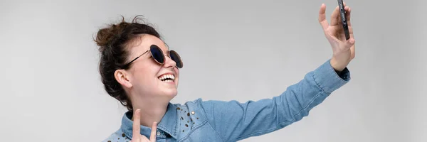 Jonge brunette meisje in ronde bril. Haren worden verzameld in een broodje. Meisje met een zwarte telefoon. Het meisje maakt selfie. — Stockfoto