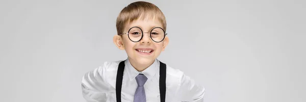 Un niño encantador con una camisa blanca, tirantes, corbata y pantalones vaqueros ligeros se levanta sobre un fondo gris. el muchacho con gafas sonríe —  Fotos de Stock