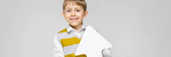 Un niño encantador con una camisa blanca, camiseta de tirantes a rayas y pantalones vaqueros ligeros está sobre un fondo gris. El niño sostiene una flecha blanca en sus manos —  Fotos de Stock