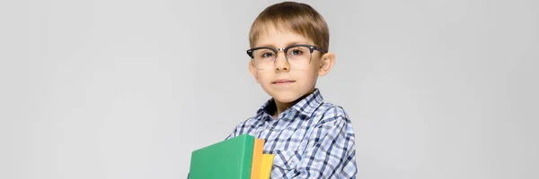 Un niño encantador con una camisa vkletchatoy y pantalones vaqueros ligeros se encuentra sobre un fondo gris. El chico tiene en sus manos una carpeta multicolor con documentos — Foto de Stock