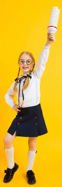 Chica con coletas rojas sobre un fondo amarillo. Una encantadora chica en gafas redondas transparentes sostiene una bombilla en sus manos . —  Fotos de Stock