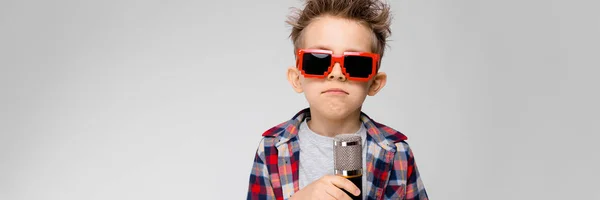 Een knappe jongen in een plaid shirt, grijs shirt en spijkerbroek staat op een grijze achtergrond. Een jongen dragen van een zonnebril. De roodharige jongen wond de draad rond zijn arm. De jongen houdt een microfoon in zijn hand — Stockfoto