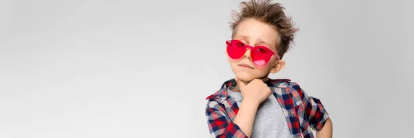 Um menino bonito em uma camisa xadrez, camisa cinza e jeans fica em um fundo cinza. Um rapaz de óculos de sol vermelhos. O menino segura uma mão em seu queixo — Fotografia de Stock