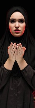 Güzel ciddi genç Müslüman kadın kavramı siyah arka plan üzerine dua olarak yüzünü yakınındaki elleriyle siyah hijab giyen portresi