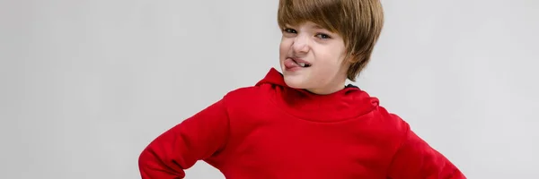 Cute confident caucasian little boy in red sweater holding hands on his waist on grey background — Stock Photo, Image