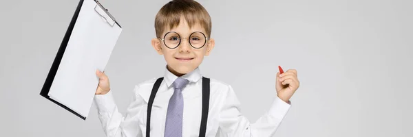 Un niño encantador con una camisa blanca, tirantes, corbata y pantalones vaqueros ligeros se levanta sobre un fondo gris. El chico sostiene un bolígrafo y sábanas para notas —  Fotos de Stock