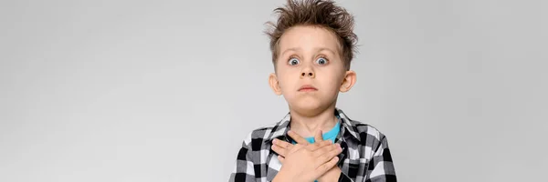 Um menino bonito em uma camisa xadrez, camisa azul e jeans fica em um fundo cinza. O rapaz dobrou as palmas das mãos no peito — Fotografia de Stock