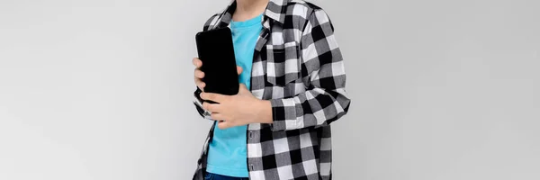 Un bel ragazzo in camicia scozzese, camicia blu e jeans si erge su uno sfondo grigio. Il ragazzo ha in mano un telefono. — Foto Stock