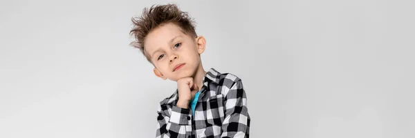 A handsome boy in a plaid shirt, blue shirt and jeans stands on a gray background. The boy supports his chin with his hand — Stock Photo, Image