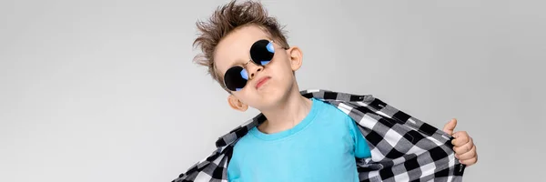 Ένα όμορφο αγόρι σε ένα καρό πουκάμισο, μπλε πουκάμισο και τζιν στέκεται σε γκρι φόντο. Το αγόρι φορώντας στρογγυλά γυαλιά. Το παιδί χαμογελάει και τραβάει το πουκάμισό του μακριά — Φωτογραφία Αρχείου