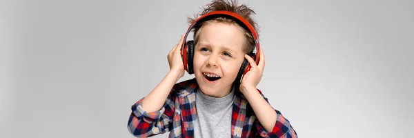 Um menino bonito em uma camisa xadrez, camisa cinza e jeans fica em um fundo cinza. Um menino de fones de ouvido vermelhos canta uma canção . — Fotografia de Stock