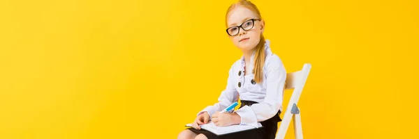 Mädchen mit roten Haaren auf gelbem Hintergrund. ein charmantes Mädchen mit durchsichtiger Brille sitzt auf einem weißen Stuhl und macht Notizen in einem Notizbuch. — Stockfoto