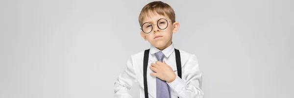 Un niño encantador con una camisa blanca, tirantes, corbata y pantalones vaqueros ligeros se levanta sobre un fondo gris. Un niño sonríe y sostiene su corbata de mano —  Fotos de Stock