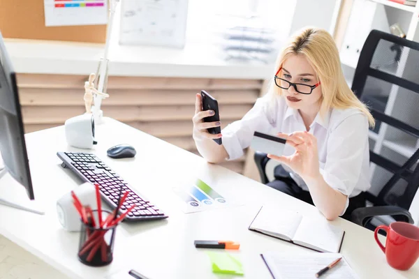 Egy fiatal lány ül az irodában, és a gazdaság egy telefon és bankkártya. — Stock Fotó