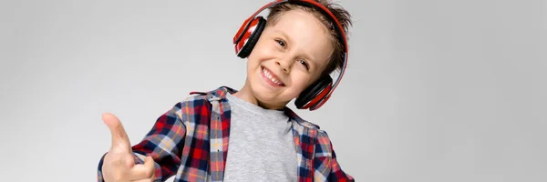 Ein gutaussehender Junge in kariertem Hemd, grauem Hemd und Jeans steht vor grauem Hintergrund. Ein Junge mit roten Kopfhörern zeigt eine Rockerziege. — Stockfoto