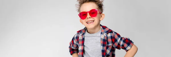 A handsome boy in a plaid shirt, gray shirt and jeans stands on a gray background. A boy in red sunglasses. The boy put his fists on his hips. The boy laughs. — Stock Photo, Image