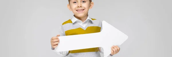 A charming boy in a white shirt, striped tank top and light jeans stands on a gray background. The boy holds a white arrow in his hands — Stock Photo, Image