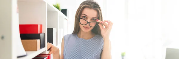 Hermosa joven en una oficina está cerca de un estante con documentos y sostiene sus gafas de mano . — Foto de Stock