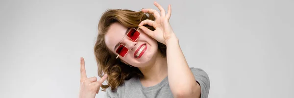 Encantadora joven con una camiseta gris sobre un fondo gris. Chica con gafas rojas de ojos cuadrados. La chica muestra una cabra rockera —  Fotos de Stock