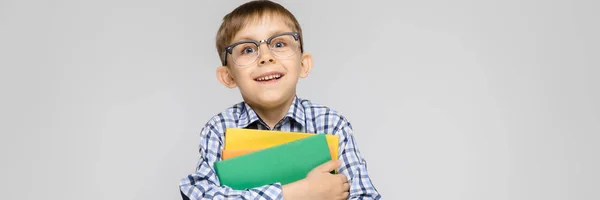 Un niño encantador con una camisa vkletchatoy y pantalones vaqueros ligeros se encuentra sobre un fondo gris. El chico tiene en sus manos una carpeta multicolor con documentos —  Fotos de Stock