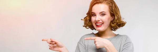 Charming young girl in a gray T-shirt on a gray background. The girl points with her fingers to the side — Stock Photo, Image